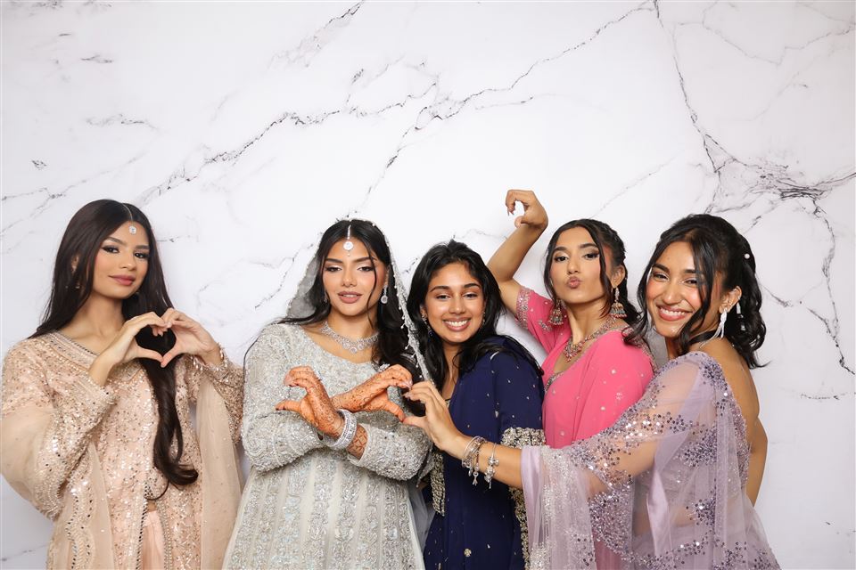 Five wedding guests in colourful formalwear make heart shapes in front of a marble photo booth backdrop