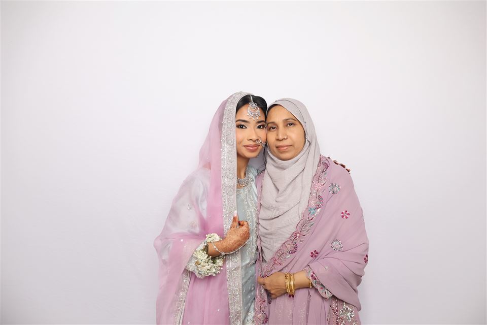 Two wedding guests in pastel formalwear pose together against a white photo booth backdrop